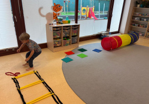 Dziecko pokonuje tor przeszkód z drabinką koordynacyjną, matami sensorycznymi i kolorowym tunelem.