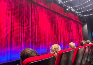 Przedszkolaki siedzą na widowni teatralnej przed sceną zasłoniętą czerwoną, kolorowo oświetloną kurtyną
