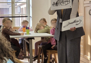 Edukatorka muzealna pokazuje przedszkolakom ilustracje zabytkowych budowli podczas warsztatów w muzeum