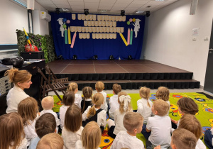 Przedszkolaki siedzą przed sceną udekorowaną z okazji Dnia Edukacji Narodowej i uroczystości pasowania na przedszkolaka.